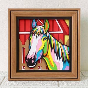 May include: A colorful papercraft horse head in a wooden frame. The horse is made of layered paper and has a pink, white, blue, green, and yellow color scheme. The horse is looking to the left and has a red barn in the background.