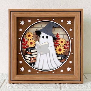Peut inclure: Un cadre photo en papier 3D avec un fantôme blanc portant un chapeau de sorcière noir et lisant un livre. Le fantôme est entouré de feuilles d'automne et de fleurs dans un cadre en bois brun.