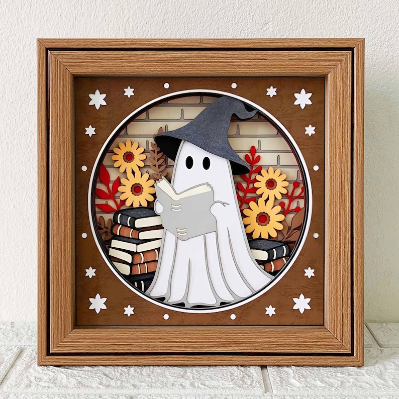 Op de afbeelding: Een 3D-papierkunst fotolijst met een witte geest die een zwarte heksenhoed draagt en een boek leest. De geest wordt omringd door herfstbladeren en bloemen in een bruin houten frame.