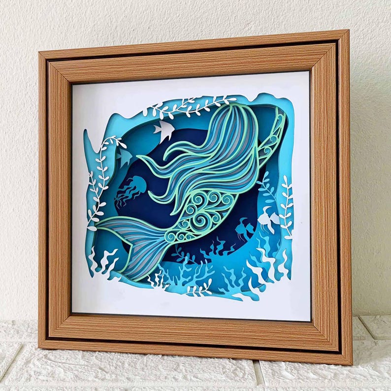 May include: A papercraft mermaid with blue and green tones, swimming through a blue and white ocean scene. The mermaid is surrounded by seaweed and jellyfish. The scene is framed in a wooden frame.