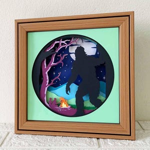 May include: A papercraft shadow box featuring a silhouette of Bigfoot walking through a forest at night. The scene is illuminated by a full moon and stars. There are three figures in the foreground around a campfire.