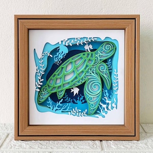 Peut inclure: Une tortue de mer découpée dans du papier vert et bleu dans un cadre en bois. La tortue est entourée d'algues et de poissons découpés dans du papier.