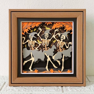 Pode incluir: Uma obra de arte em papel com tema de Halloween emoldurada. A obra apresenta três esqueletos dançando com chapéus de bruxa e saias cinzentas. O fundo inclui árvores laranja e um fundo preto, tudo dentro de uma moldura de madeira castanha.