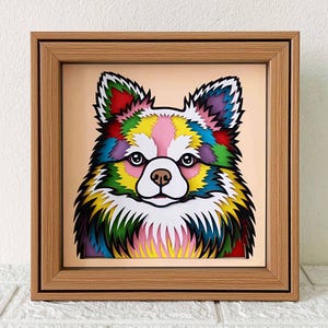 May include: A framed paper art piece of a colorful dog with a white face and fluffy fur. The dog is surrounded by a rainbow of colors, including red, orange, yellow, green, blue, indigo, and violet. The frame is made of wood and has a light brown finish.