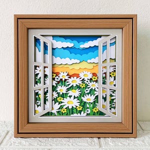 May include: A framed paper art piece showing a sunny outdoor scene. The artwork features a window with open shutters, revealing a field of white daisies and a colourful sky with blue, yellow, and white layers. The frame is made of wood.