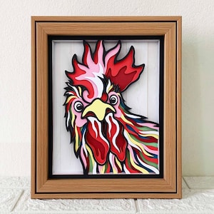 Könnte beinhalten: Ein bunter Papier-Hahn mit einem roten, gelben und grünen Kamm und Kehllappen. Der Hahn ist in einem Holzrahmen mit schwarzem Passepartout gerahmt.