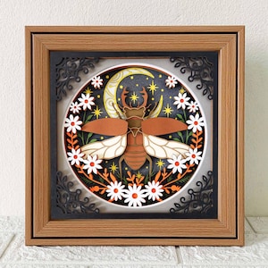 May include: A framed paper art piece featuring a brown beetle with white and brown wings, set against a night sky with a crescent moon, stars, and white daisies. The frame is brown with a black filigree border.