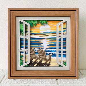 May include: A framed paper art piece depicting a beach scene viewed through an open window. The artwork features a sunset over the ocean, two beach chairs, and palm trees. The frame is made of light brown wood.
