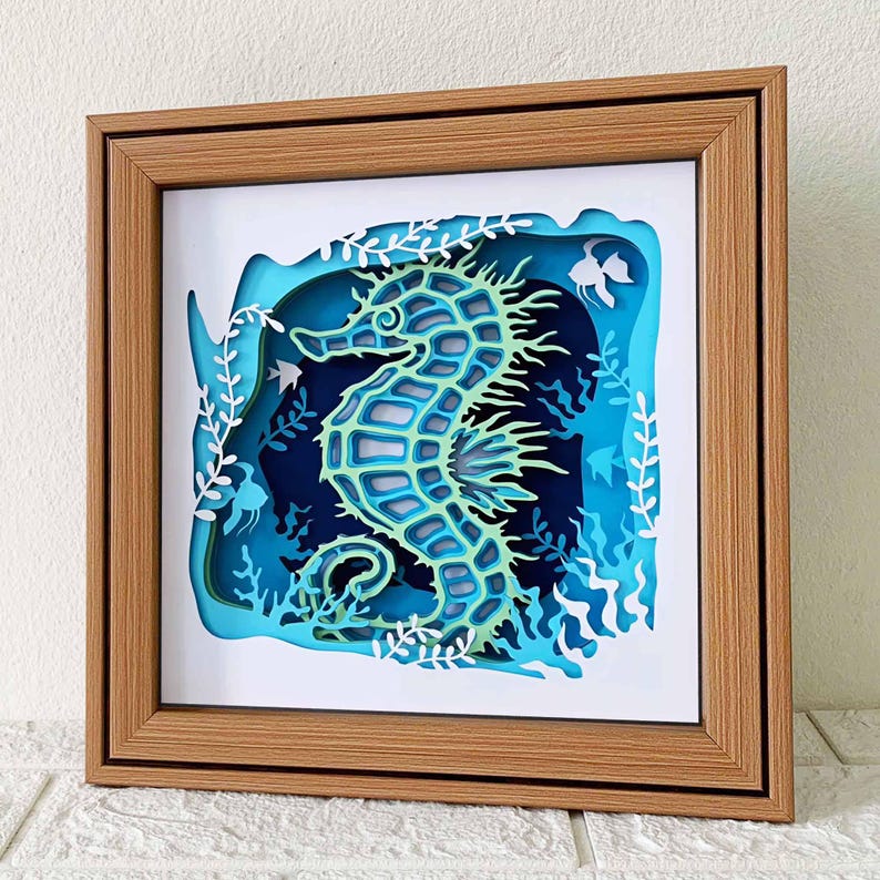 May include: A wooden frame with a blue and green paper cut-out of a seahorse surrounded by seaweed. The seahorse is in a 3D style and appears to be swimming in the ocean.