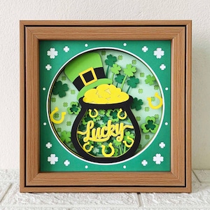 May include: A framed St. Patrick's Day paper art piece. The artwork features a green leprechaun hat, a pot of gold, shamrocks, and the word "Lucky" in yellow script. The background is green with white accents, and the frame is brown.