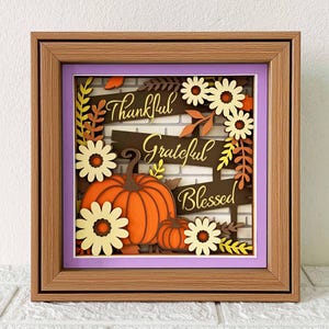 May include: A wooden frame with a layered paper design featuring pumpkins, flowers, and leaves. The words "Thankful", "Grateful", and "Blessed" are written in gold lettering on the frame.
