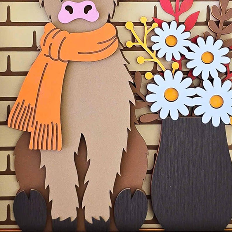 May include: A layered paper art piece featuring a Highland cow wearing an orange scarf, with a vase of white daisies. The cow is brown and tan, set against a brick-like background. The flowers have yellow centers.