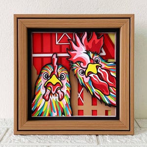 Könnte beinhalten: Ein gerahmtes Papierbastelwerk mit zwei bunten Hühnern mit roten, gelben und blauen Akzenten. Die Hühner schauen hinter einer roten und weißen Scheunenstruktur hervor.