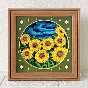 Może przedstawiać: Ramka z papierową grafiką 3D przedstawiającą grupę słoneczników na tle niebieskiego i białego nieba. Słoneczniki są żółte z brązowymi środkami i zielonymi liśćmi. Ramka jest wykonana z brązowego drewna z białym passe-partout.