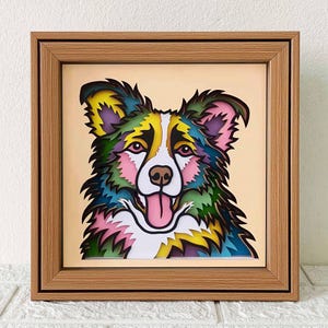 Op de afbeelding: Een ingelijst kunstwerk met een kleurrijke, gelaagde papiersnijkunst van een hondenkop. De hond heeft een wit gezicht met roze wangen, een zwarte neus en een roze tong die uitsteekt. De vacht van de hond is een mix van groen, blauw, geel en roze.