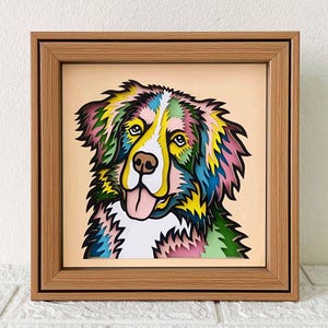 May include: A colorful, layered paper art piece of a Bernese Mountain Dog with a pink tongue sticking out. The dog is framed in a light brown wood frame.