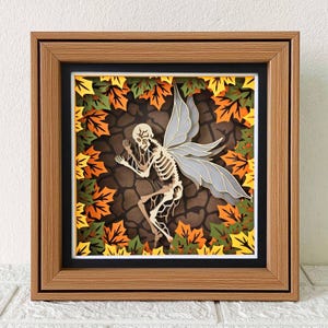 May include: A framed paper art piece depicting a skeleton fairy with grey wings, surrounded by autumn leaves in orange, yellow, and green. The artwork is set against a brown background and framed in a wooden frame.