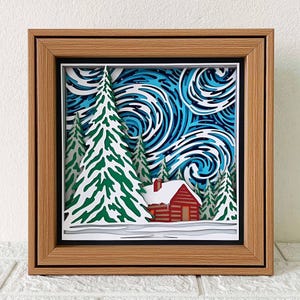 May include: A papercraft 3D art piece depicting a snowy winter scene with a red cabin, pine trees, and a swirling blue sky. The artwork is framed in a wooden frame.