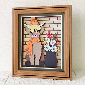 May include: A framed 3D paper art piece featuring a Highland cow wearing an orange hat and scarf, set against a brick wall backdrop. A vase of flowers sits beside the cow. The artwork is encased in a brown wooden frame.