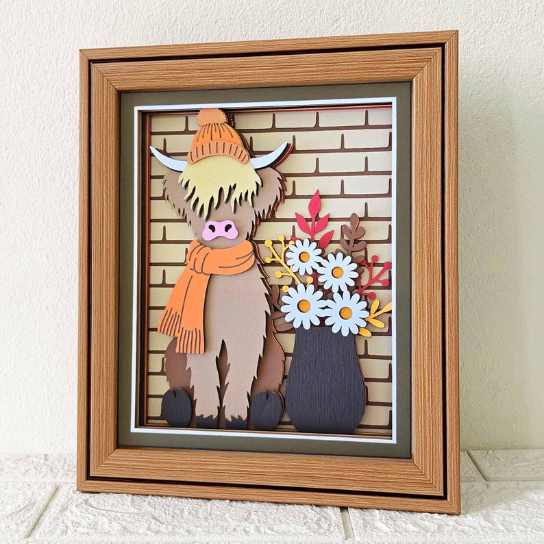 May include: A framed 3D paper art piece featuring a Highland cow wearing an orange hat and scarf, set against a brick wall backdrop. A vase of flowers sits beside the cow. The artwork is encased in a brown wooden frame.