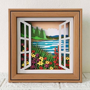 May include: A papercraft window scene with a view of a lake and forest. The window is open and there are flowers in the foreground. The scene is framed in a wooden frame.