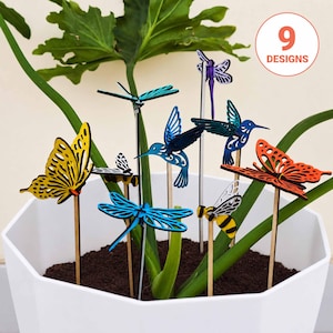 May include: A collection of nine decorative plant stakes featuring laser-cut wooden figures of butterflies, dragonflies, hummingbirds, and bees in vibrant colors like yellow, blue, orange, and purple, all arranged in a white hexagonal planter.