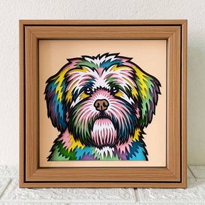 May include: A colorful paper cut-out of a dog's face in a wooden frame. The dog has a pink nose, black eyes, and a white mouth. The paper cut-out is layered with different colors, including green, blue, yellow, and pink.