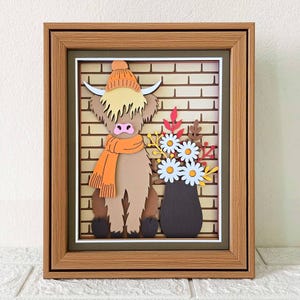 May include: A framed paper art piece featuring a Highland cow wearing an orange hat and scarf, set against a brick wall backdrop. A vase of white daisies and fall foliage sits beside the cow. The frame is a light brown color.