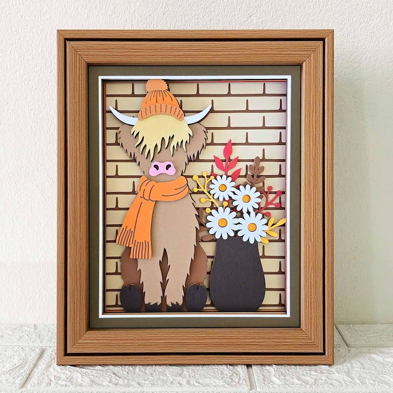 May include: A framed paper art piece featuring a Highland cow wearing an orange hat and scarf, set against a brick wall backdrop. A vase of white daisies and fall foliage sits beside the cow. The frame is a light brown color.