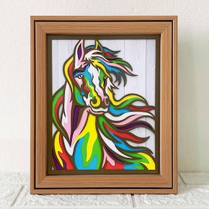 Peut inclure: Une découpe en bois de tête de cheval colorée dans un cadre brun. La crinière du cheval flotte au vent et est composée de différentes couleurs, dont le rose, le violet, le bleu, le vert, le jaune et le blanc.