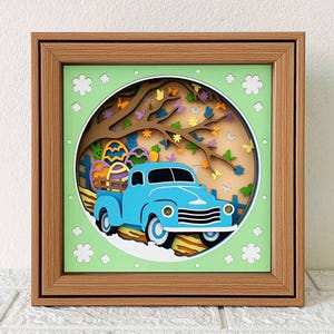 May include: A blue pickup truck with a wooden bed filled with colorful Easter eggs drives through a green, paper-cut forest scene. The truck is framed in a brown wooden frame with a green mat.