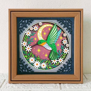 May include: A framed, layered paper art piece featuring a green and orange hummingbird with white and green wings, surrounded by white daisies and a crescent moon. The background is a pinkish-purple with yellow stars. The frame is a light brown color.