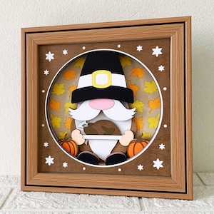 May include: Thanksgiving-themed framed artwork featuring a gnome wearing a pilgrim hat with a gold buckle. The gnome holds a turkey, with pumpkins on either side. The background has fall leaves and stars, all within a wooden frame.