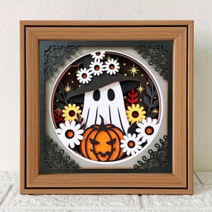 Può includere: Un'opera d'arte su carta a tema Halloween incorniciata. L'opera presenta un fantasma bianco con un cappello da strega nero, una zucca arancione intagliata e fiori bianchi e gialli. La cornice è marrone con angoli decorativi neri.