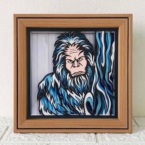 Può includere: Un'opera d'arte in cartapesta incorniciata che raffigura un Bigfoot blu e bianco con un'espressione seria. L'opera d'arte è posizionata su uno sfondo a grana di legno e uno sfondo a strisce blu e bianche.