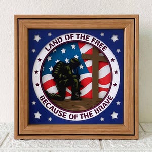 Puede incluir: Una obra de arte enmarcada con temática patriótica, que representa la silueta de un soldado arrodillado frente a una bandera estadounidense y una cruz. El diseño incluye el texto "LAND OF THE FREE" y "BECAUSE OF THE BRAVE" en una disposición circular, sobre un fondo azul con estrellas blancas.