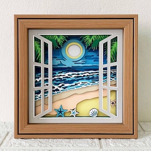 Puede incluir: Un marco de madera con un diseño de papel recortado de una ventana abierta que mira hacia una escena de playa nocturna. La escena incluye una luna llena, olas del océano, arena, palmeras y conchas marinas.