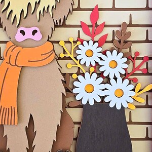 May include: A layered paper art piece featuring a Highland cow wearing an orange scarf, and a vase of white daisies with yellow centers. The background is a brick wall, with autumn-colored leaves.