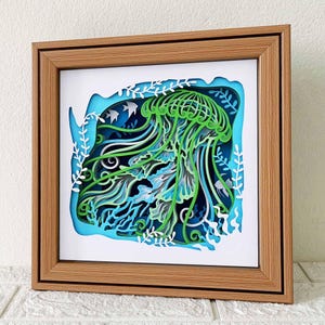 May include: A wooden frame with a 3D paper cut-out of a green jellyfish with blue and white accents. The jellyfish is surrounded by white paper cut-out seaweed and coral.