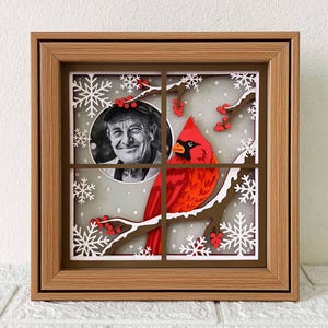 May include: A framed papercraft artwork featuring a red cardinal perched on a branch with snowflakes and red berries. A black and white photo of a man is in the background.