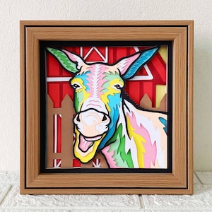 May include: A colorful paper cutout of a donkey's head with a red barn and fence in the background. The donkey is smiling with its mouth open and tongue sticking out. The donkey is made of paper and has a layered, 3D effect.