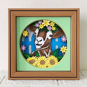 May include: A framed paper art piece featuring a brown goat with a floral crown, set against a light green background. The goat is in front of a blue picket fence, surrounded by yellow sunflowers and other colorful flowers.