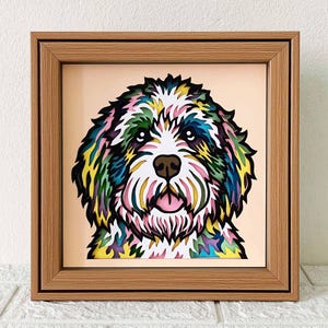 May include: A framed paper art piece of a dog's face. The dog is a white, fluffy breed with a pink tongue and black nose. The dog's fur is layered in shades of green, blue, yellow, and pink.
