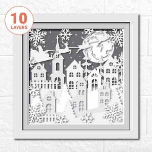 May include: A white paper cut-out design of a winter village scene with Santa Claus and reindeer flying over a full moon. The design is framed in white and has 10 layers.