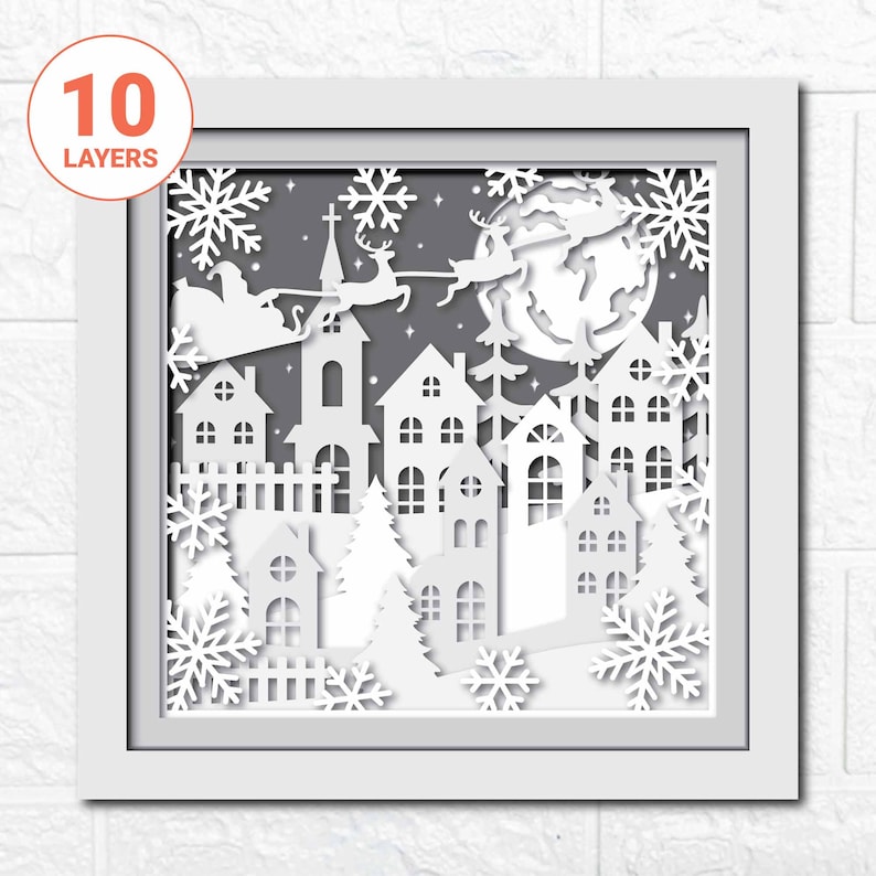 May include: A white paper cut-out design of a winter village scene with Santa Claus and reindeer flying over a full moon. The design is framed in white and has 10 layers.