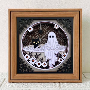 Könnte beinhalten: Gerahmtes Halloween-Papierkunstwerk. Das Kunstwerk zeigt einen Geist in einer Badewanne mit einer schwarzen Katze, umgeben von Blasen, Gänseblümchen und einem Spinnennetz. Der Rahmen ist braun mit einem schwarzen Filigranrand.