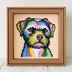 Op de afbeelding: Een ingelijst, gelaagd papierkunstwerk met een kleurrijk hondenportret. De vacht van de hond is afgebeeld in tinten blauw, groen, geel, roze en paars. Het kunstwerk staat tegen een lichtperzik achtergrond en is ingelijst in een houten lijst.