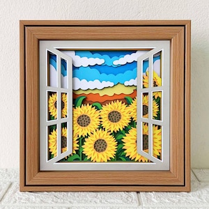 Może przedstawiać: Oprawione dzieło sztuki z papieru przedstawiające okno z widokiem na pole słoneczników. Białe okno z otwartymi okiennicami ukazuje warstwową scenę słoneczników, niebieskiego nieba i chmur. Rama jest w kolorze jasnobrązowym.