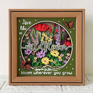 May include: A framed, layered paper art piece featuring a wildflower theme. The artwork includes colorful paper flowers, butterflies, and the text "Live like a wildflower, bloom wherever you grow." The frame is a light brown color.