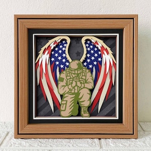 May include: A framed layered paper art piece featuring a kneeling soldier with angel wings. The wings display the American flag design in red, white, and blue. The artwork is set against a black background with stars and stripes, encased in a wooden frame.